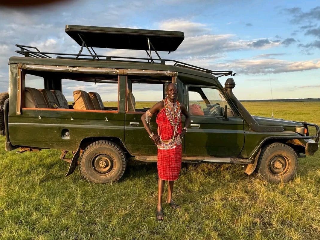Jackson Karia, Maasai driver-guide, standing beside an open-roof 4x4 Land Cruiser on the Maasai Mara